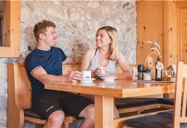 Ein Paar sitzt an einem Holztisch in einer gemütlichen Umgebung. Sie lächeln und trinken Kaffee zusammen.