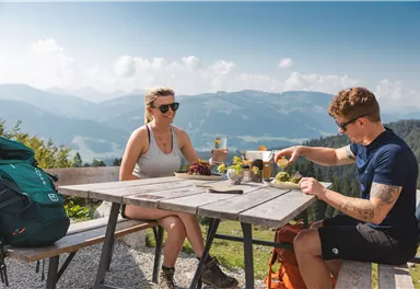 Ein Paar sitzt an einem Tisch im Freien und genießt eine Mahlzeit in den Bergen. Die Aussicht zeigt eine schöne Landschaft mit Bergen und blauem Himmel.