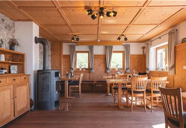 Ein gemütlicher Raum mit Holzmöbeln und einem Holzofen. Große Fenster lassen viel Licht herein und schaffen eine einladende Atmosphäre.