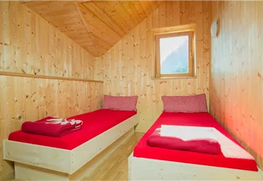 Ein gemütliches Zimmer mit zwei Einzelbetten und warmen roten Bettwäsche. Die Wände sind aus Holz, und ein kleines Fenster lässt Tageslicht herein.