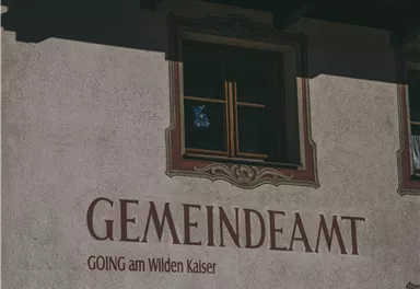The image shows the facade of a building with the inscription "Municipality" and "Going am Wilden Kaiser." A window is visible, and the walls are simply designed.