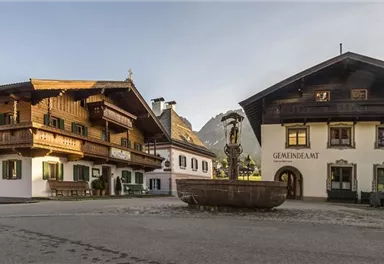 Ein malerischer Dorfplatz mit traditionellen Holzgebäuden. In der Mitte befindet sich ein schöner Brunnen, umgeben von einer ruhigen Landschaft.