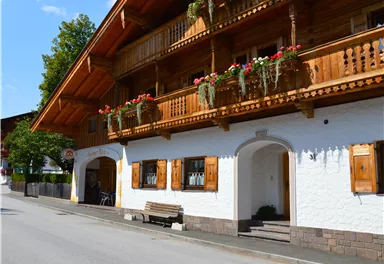A traditional chalet with wooden balustrades and flower boxes. The street in front of the building is quiet and lined with trees.