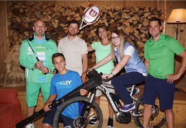 Eine Gruppe von sechs sportlichen Personen posiert in einem gemütlichen Raum. Sie tragen Freizeitkleidung und halten verschiedene Sportgeräte wie ein Fahrrad und einen Tennisschläger.