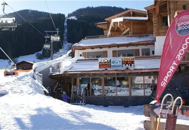 Ein Schneegebiet mit einem Bergrestaurant und Skiausleihe. Im Hintergrund sind Skilifte und schneebedeckte Berge zu sehen.