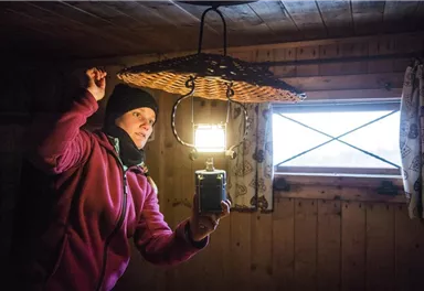 A person stands in a wooden room and holds a lamp over a basket. It is dark, but the light from the lamp illuminates the room.
