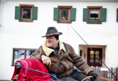 A smiling older man sits on a carriage. In the background, there is a traditional house with green shutters.