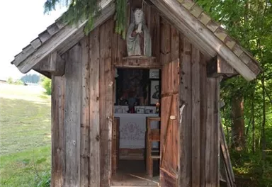 A small wooden cabin stands surrounded by trees in a meadow. The door is open and reveals a room with a religious statue.