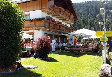 A cozy restaurant with umbrellas and guests outside, surrounded by green meadows and mountains. The facade of the house is decorated in a traditional style with flower boxes.