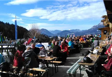 A lively terrace with many guests enjoying the sun. In the background, mountains and a clear sky can be seen.