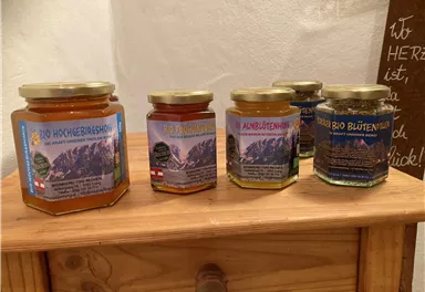 Various jars of honey are on a wooden table. Each jar has a label featuring a mountain landscape and descriptions in German.