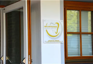 Ein Eingang mit einer modernen Beschilderung für eine Zahnklinik. Die Glasfront und das Holzrahmenfenster schaffen eine einladende Atmosphäre.