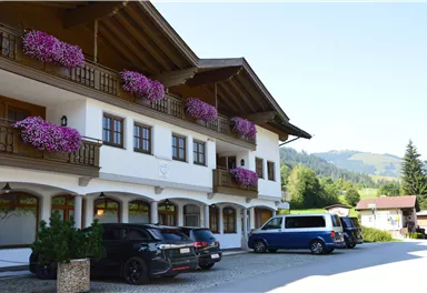 Ein malerisches Gebäude im bayerischen Stil mit Balkonblumen. Im Vordergrund parken mehrere Autos vor einer schönen Berglandschaft.