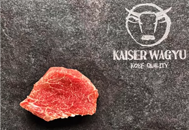 A raw piece of Wagyu meat on a dark surface. Next to it is the "Kaiser Wagyu" logo for the highest quality.