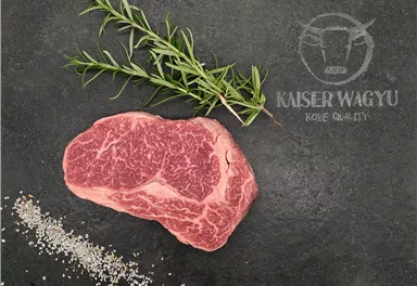A high-quality piece of Wagyu beef lies on a dark background. Next to it are fresh herbs and salt sprinkles.