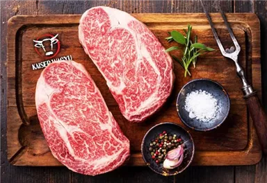 Zwei hochwertige Steakstücke auf einem Holzbrett, umgeben von Gewürzen und frischen Kräutern. Im Hintergrund ist das Logo „KAISERWAGYU“ sichtbar.