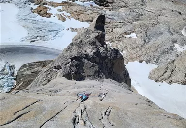 A climber ascends a steep rock face with a view of an impressive mountain landscape. Snow and glaciers are visible in the surrounding area.