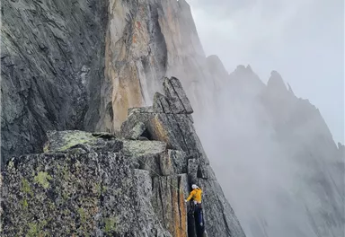 A climber ascends a steep, rocky peak with fog in the surroundings. The dramatic landscape showcases tall rocks and a mystical atmosphere.