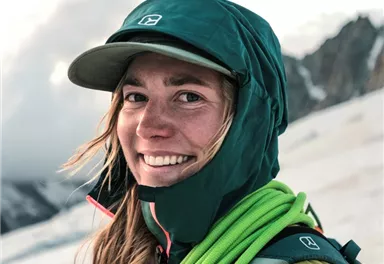 A smiling person with a green hat and weatherproof clothing stands in the mountains. In the background, clouds and mountain landscapes can be seen.