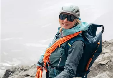 A climber stands on a rock, wearing a helmet and equipment. She smiles at the camera and appears ready for the adventure in the mountainous landscape.