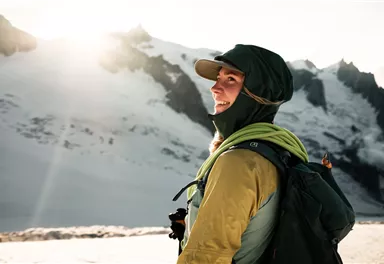 A person stands on the mountain and smiles into the distance. The landscape is covered in snow and the sun is shining brightly.