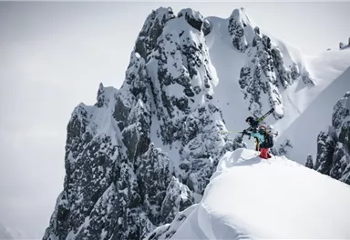 A skier stands on a snow-covered summit with impressive rocks in the background. The scene conveys a sense of adventure in a winter mountain landscape.