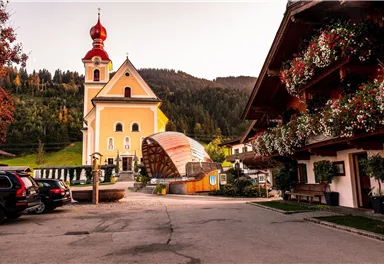 A picturesque village street with a church and traditional wooden buildings. In the background, wooded hills are visible.
