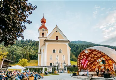 A picturesque church with a red tower stands in the background. In front of the church, a concert is taking place in a wooden pavilion, surrounded by an audience and green nature.