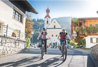 Two women are riding their bicycles through a picturesque village. In the background, there is a church, and the surroundings are bordered by mountains.