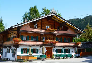 A traditional wooden house in alpine style with green shutters and blooming balconies. It is located surrounded by trees and a clear blue sky.