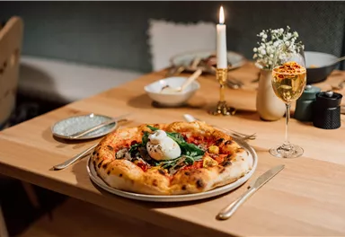 Ein Tisch mit einer köstlichen Pizza, belegt mit frischem Gemüse und einem Ei. Daneben steht ein Glas Champagner und eine Kerze sorgt für eine gemütliche Atmosphäre.