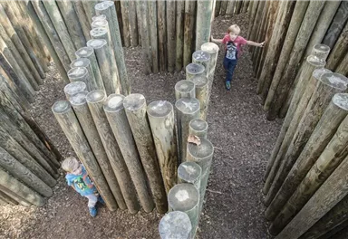 A playground maze made of wooden logs with two children playing inside. The surroundings are natural and inviting.