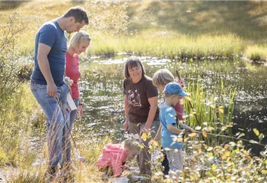 A family is standing at the edge of a pond and fishing. Children are playing in the water and adults are watching them.