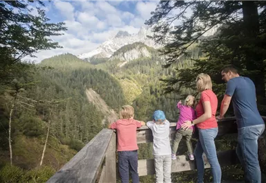 A family stands on a viewing platform and gazes at an impressive mountain landscape. In the background, green forests and snow-covered peaks are visible.