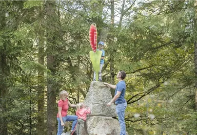 A family is climbing a large rock in the forest. A child is standing on top of the rock and holding a colorful sign.