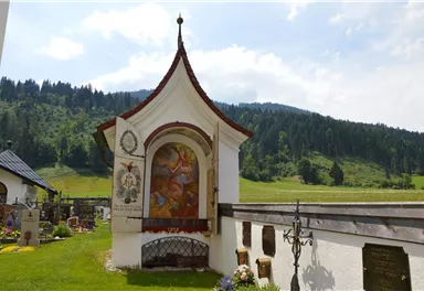 Ein elegantes Gotteshaus mit bunten Fresken steht in der grünen Landschaft. Im Hintergrund sind sanfte Hügel und ein blauer Himmel zu sehen.