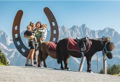 Eine Familie steht glücklich vor einem Hoftor aus Hufeisen. Im Hintergrund sind Berge zu sehen, und neben ihnen steht ein kleiner Pony.