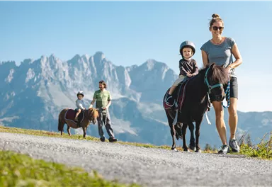 Eine Frau führt ein Pony mit einem kleinen Kind auf dem Rücken. Im Hintergrund sind Berge und eine zweite Familie zu sehen.