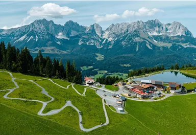Eine malerische Landschaft mit grünen Wiesen und majestätischen Bergen im Hintergrund. Im Vordergrund liegt eine kleine Siedlung mit einem Teich und gewundenen Wegen.