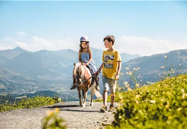 Ein Mädchen reitet auf einem Pony, während ein Junge es auf einem Weg in einer bergigen Landschaft führt. Der Himmel ist blau und die Umgebung ist grün und blühend.