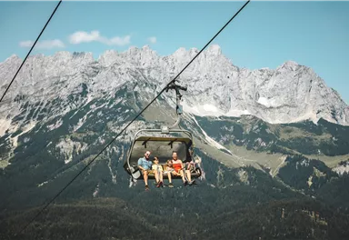Eine Seilbahn mit vier Personen fährt über eine wunderschöne Berglandschaft. Im Hintergrund sind majestätische Berge und ein blauer Himmel zu sehen.