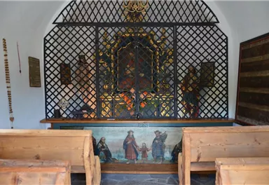 A small chapel with wooden benches and an intricately decorated altar. The walls are plain, and the windows are fitted with a grille.