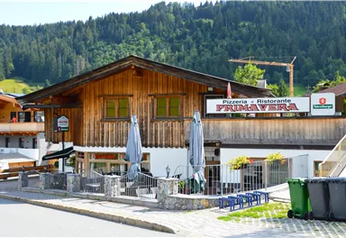 A cozy pizzeria with a wooden facade and outdoor terrace. Surrounded by green mountains and a quiet street.