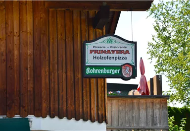A restaurant sign with the name "Primavera" for wood-fired pizza. The background features wooden cladding and green trees.