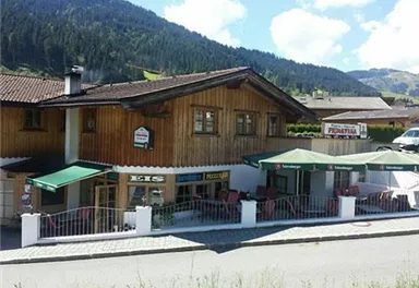 A traditional wooden house with a restaurant. Outside there are tables with umbrellas and in the background, green mountains can be seen.