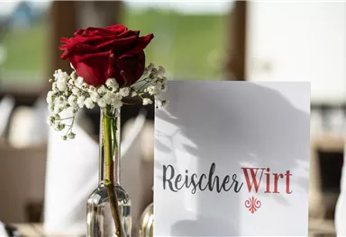 Eine elegante Tischdekoration mit einer roten Rose in einer Glasvase und einer Menükarte. Im Hintergrund ist eine grüne Landschaft zu sehen.
