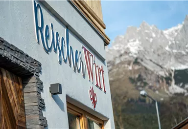 Ein Gebäude mit dem Schriftzug "ReiseWirt" in einer malerischen Berglandschaft. Die Umgebung ist bergig und zeigt schneebedeckte Gipfel im Hintergrund.