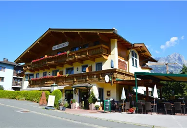 Ein charmantes alpines Hotel mit Holzverzierung und Balkonen. Im Hintergrund sind schöne Berge und ein klarer blauer Himmel zu sehen.