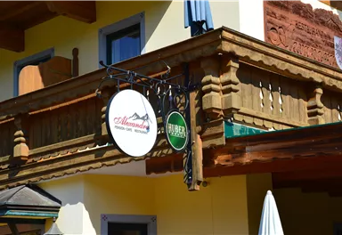 Ein traditionelles Gebäude mit einem Holzbalkon. Davor hängen zwei Schilder, eines mit dem Namen "Alpenblick" und das andere mit dem Hinweis auf Bier.