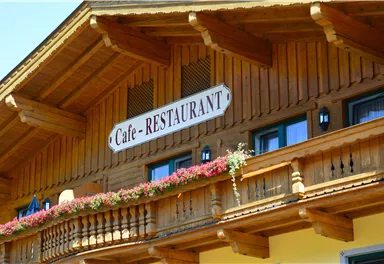 Ein traditionelles Holzgebäude mit einem Schild, das "Cafe - RESTAURANT" zeigt. Auf dem Balkon blühen bunte Blumen und die Fenster sind klein und einladend.